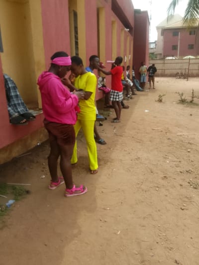 The image depicts an outdoor scene in Umuokpu, Nigeria, adjacent to a building with reddish-pink walls and vertical yellow-ochre accents. The ground consists of reddish-brown dirt. In the foreground, two individuals are standing. The person on the left wears a hot pink hooded sweatshirt, brown shorts, a pink headband, and pink sandals; they are looking down. The person on the right wears a bright yellow top and matching trousers with brown sandals; they are also looking down and appear to be interacting closely with the first individual, their hands near each other. A black and white plaid garment is draped over a low ledge to the far left.
Further along the building's wall, several other individuals are present. One person in a red t-shirt and black-and-white checkered shorts stands, holding a pink cylindrical object near another person's face. Other figures are seated or standing along the wall, some appearing to rest. In the background, a brown metal fence separates the dirt area from a multi-story brown building with a light-colored roof. Sparse green vegetation is visible on the dirt ground and a palm frond extends into the upper right corner.