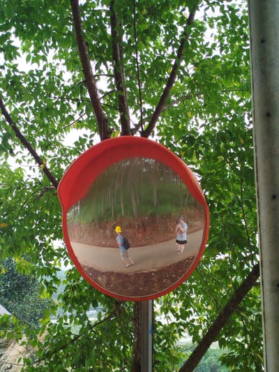 A convex mirror, mounted on a pole and framed in red, reflects a street scene. The mirror is positioned outdoors amidst lush green foliage and tree branches. In the reflection, a person wearing a yellow hard hat and a blue shirt with a black backpack is walking away from the viewer on a paved road. To the right, another person is reflected, wearing a gray t-shirt, dark skirt, and sandals, and is holding a phone, appearing to take a photograph. The road in the reflection is lined with trees and a grassy embankment. The surrounding environment suggests a slightly rural or suburban setting within the Cam Ly area of Da Lat, Vietnam, characterized by verdant hillsides and a winding road.
