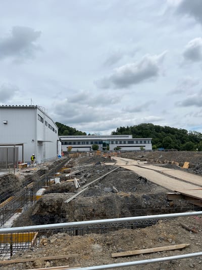 The image depicts an outdoor construction site in Hanno, Japan, under a cloudy sky. In the foreground, deep trenches are dug, revealing rebar frameworks for concrete foundations. Construction materials, including wooden planks and metal pipes, are scattered around the trenches. A person wearing a yellow vest and a white hard hat stands near a building on the left side of the image. This building is a modern, silver-colored structure with multiple windows. Further in the background, another long, multi-story building with a white facade and dark windows is visible. Behind the buildings, a dense, green forested hill rises. The overall scene conveys ongoing construction work in a semi-urban or suburban environment.
