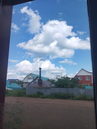 This media file depicts a daytime outdoor scene, likely in a rural or suburban area of Birsk, Russia, given the provided location context. The sky is a vibrant blue, dotted with large, fluffy white cumulus clouds, suggesting fair weather.
In the foreground, a dirt road or path is visible, with some sparse vegetation growing along its edge. Through what appears to be a window or frame, several houses can be seen. One prominent house has a turquoise roof and light-colored siding, with a brick chimney. Next to it is another house with a darker, possibly brown, roof and a tall, dark chimney. Further to the right, a larger brick house with white trim and a white gable is visible. Trees and green foliage partially obscure some of the structures. A grey metal fence runs along the property line, and some graffiti, including a heart symbol, is present on it. The overall impression is a peaceful, residential setting on a pleasant day. No people are visible in the frame.