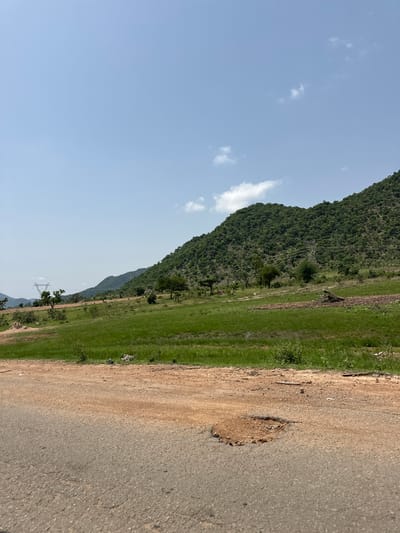 The image depicts a rural landscape in Lankoviri, Nigeria. A paved road with a prominent pothole is in the foreground, with a dirt shoulder bordering it. Beyond the road, a vast expanse of lush green grassland stretches towards a densely forested hillside. In the mid-ground, scattered trees dot the landscape, and a tall electricity pylon is visible on the left. The sky is a clear, pale blue with a few wispy white clouds. The scene is characterized by natural beauty and hints of rural infrastructure.