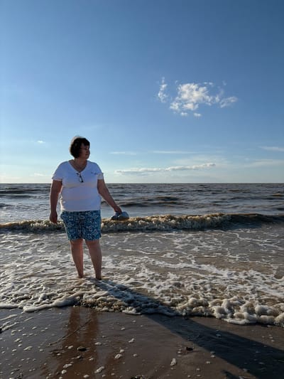 In the city of Port-Katon, Russia, a woman stands in shallow ocean water under a blue sky with a single cloud. She is wearing a white t-shirt and blue patterned shorts, and is holding a pair of light-colored shoes in her right hand. The water is slightly choppy with small waves, and the shoreline is sandy with some foam.