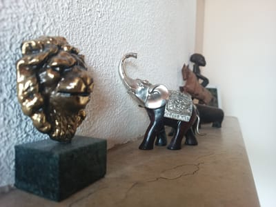The image presents a shelf with several decorative objects. On the left is a gold-colored lion head sculpture mounted on a dark green marble or similar stone base. To the right of the lion head is a silver and dark brown elephant figurine with an ornate saddle cloth. Further back, there is a wooden horse and rider sculpture and another dark-colored animal figurine, possibly a hippopotamus. The shelf appears to be made of a light brown, faux-marble material. The background consists of a textured, off-white wall and a glimpse of a plain white wall in the upper right corner. The location is not discernible from the visual information provided.