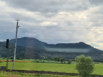 A wide-angle shot captures a pastoral landscape with a focus on a mountain range under a cloudy sky. In the foreground, a railway track runs horizontally across the image, bordered by tall grasses on the left and a vibrant green field on the right. A solitary tree with dense foliage stands prominently in the middle ground, to the right of the frame. Behind the fields, a fence line is visible, separating the green fields from a patch of orange-colored land. The background is dominated by a large mountain, partially shrouded in a misty haze, with its slopes covered in dark green trees. Several power lines stretch across the upper portion of the image, supported by a tall, slender pole on the left, which also features a railway signal. The sky is overcast with thick, grey clouds, punctuated by lighter patches suggesting diffused sunlight. The scene evokes a sense of serene rural tranquility, likely captured from a moving vehicle.