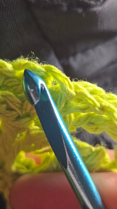 A close-up photograph shows a teal-colored crochet hook in the process of working a stitch with bright green yarn. The yarn appears to be fluffy and somewhat loosely spun. A portion of a hand can be seen holding the hook. The background is out of focus and consists of a dark gray material, possibly clothing or a carrying bag. The image does not offer information about the location. Thus, it is not possible to determine if it was taken in Lompoc, United States of America.