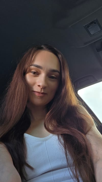 A young woman with long, wavy, brown hair is captured in a close-up, slightly angled shot. She is wearing a white ribbed tank top and appears to be inside a vehicle, as indicated by the dark interior lining and a window visible on the right side of the frame. The lighting suggests she is outdoors, possibly during the daytime, with sunlight catching her hair and casting subtle shadows on her face. Her expression is a gentle smile, with her gaze directed slightly off-camera. The background is dark and out of focus, providing no specific environmental details of Bulgakovo, Russia.
