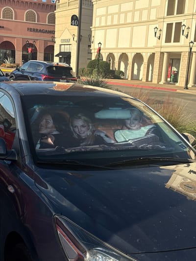 Three smiling women are seen inside a dark car through the windshield. The reflections in the windshield show buildings with retail stores like Ferragamo and Citizen. The car is dark blue and parked on a sunny day. Other cars are parked nearby. The scene suggests a shopping area with multiple storefronts. The light and reflections add depth to the composition.