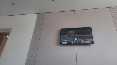 In an indoor setting, a television screen displays a movie trailer. The screen shows a woman with blonde hair looking to the right, and below her, a smaller image of a man's face is visible. Text on the screen reads "ОНИ ДУМАЛИ, ЧТО АКУЛЫ БЫВАЮТ ТОЛЬКО В ОКЕАНЕ | Захватывающая хоррор" and "ТАЙГА". The television is mounted on a light-colored wall with vertical textured panels on the left and right sides. The environment appears to be a modern, sparsely decorated interior space. This scene is located in Қаражар, Kazakhstan.