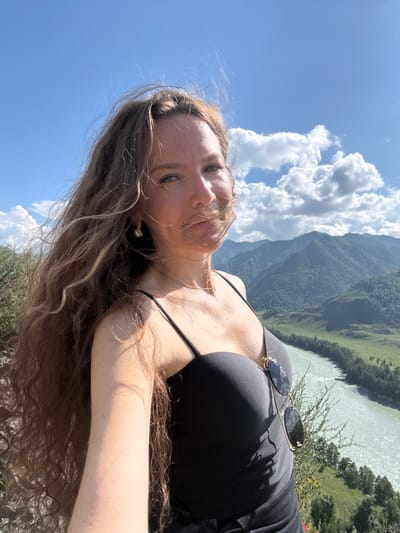 A woman with long, wavy brown hair is taking a selfie on a sunny day with a scenic mountain and river backdrop. She is wearing a black tank top with spaghetti straps and has sunglasses clipped to the neckline of her top. Her hair is blowing in the wind. In the background, lush green mountains rise up, and a wide, turquoise river flows through a valley. The sky is blue with scattered white clouds. The location is a mountain overlook near the Katun River in Chemal, Russia.