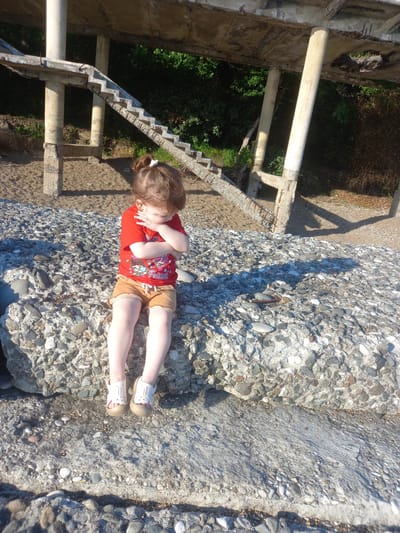 A young girl with brown hair tied in a bun is sitting on a large concrete structure composed of many rocks. She is wearing a red t-shirt with graphics and tan shorts, and white sandals. Her arms are crossed and she is looking down. The concrete structure is on a beach, with sand and smaller pebbles around it. In the background, there is a partially ruined concrete structure with columns and stairs, surrounded by green foliage. The overall setting appears to be an outdoor, somewhat neglected, coastal area. This location is identified as Gwandra, Abkhazia.