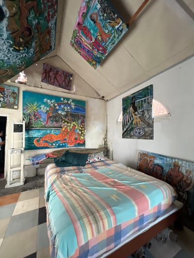 This is a full-shot, eye-level, interior photograph of a bedroom in Tejakula, Indonesia. The room is filled with various paintings and artworks, suggesting it might be an artist's studio or a space dedicated to showcasing art. A prominent feature is a large bed with a colorful, striped bedspread that covers most of the foreground. The bed is positioned at an angle, with its foot towards the right side of the frame.
On the wall behind the bed, there are several paintings. To the left of the bed, a large, vibrant painting depicts a tropical scene with a mermaid-like figure and a jaguar. Above and to the left, another painting with figures is visible, as well as a smaller horizontal painting with dancing figures. The ceiling also has artwork, with two paintings mounted on it, one featuring abstract figures and the other a more detailed scene with figures and vehicles.
To the right of the large tropical painting, another painting hangs on the wall, showing a person sitting on a balcony with a drink and a plate of food. Further to the right, on the floor next to the bed, two more paintings are leaning against a wall, one depicting faces and another with figures.
A white ladder-like structure with a sign that reads "ART ZOO" is visible to the left of the bed, near an open doorway. The floor is tiled with a geometric pattern of squares in various shades of grey, brown, and orange. The walls are white and somewhat textured, with some signs of age or wear. The overall impression is a bohemian and art-filled living space.