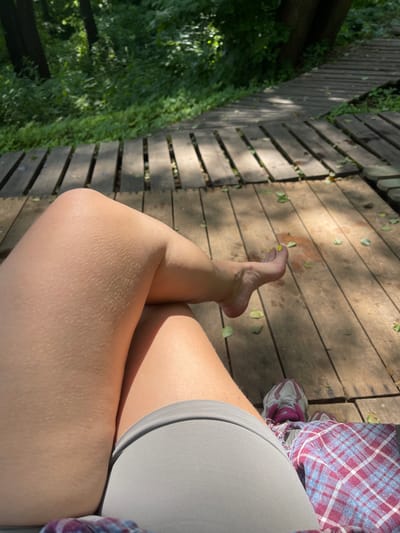 The image shows a person's legs resting on a wooden boardwalk in a natural, outdoor setting. The boardwalk is constructed of weathered wooden planks and is surrounded by lush green foliage, suggesting a park or wooded area. Sunlight filters through the leaves, creating dappled shadows on the boardwalk. The person is wearing gray shorts and a plaid shirt. One foot, with yellow toenail polish, is visible, resting on the boardwalk. A pair of athletic shoes is also visible near the person's feet. The scene appears to be a moment of relaxation in a natural environment.