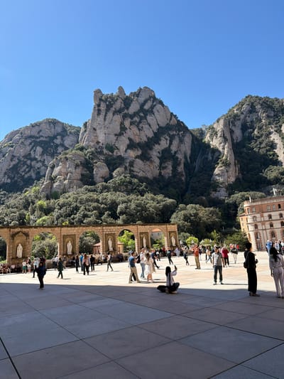 This image captures a bright, sunny day at the Montserrat complex in Monistrol de Montserrat, Spain, showcasing a blend of natural beauty and architectural heritage.
The scene is dominated by the majestic, rugged mountains of Montserrat, visible on the left and extending into the background. These distinctive rocky formations are partially covered in lush green vegetation. A clear, vibrant blue sky fills the upper portion of the frame. A faint funicular track can be observed ascending one of the mountain slopes in the distance, providing a key location cue.
In the mid-ground, a grand architectural structure stands on the right. This multi-storied building features a warm-toned facade with numerous arched windows. Below it, a series of ornate stone arcades stretches horizontally, each arch housing a white statue, likely depicting religious figures.
The foreground and right side of the image are occupied by a spacious, light-colored paved plaza. Numerous people, comprising adults and possibly some children, are scattered across this area. Many appear to be tourists or visitors, walking casually, standing and observing their surroundings, or gathering in what resembles a queue along the right edge of the plaza, partially shaded by the building.
The bright sunlight casts distinct, elongated shadows across the plaza, indicating a mid-morning or early afternoon time of day. The weather is clear and pleasant. No discernible text is visible anywhere in the image.