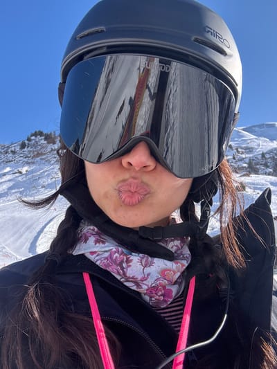 A close-up image depicts an individual on a ski slope in Zürs, Austria. The person is wearing a black Giro ski helmet and large, black mirrored ski goggles branded "SUNDOGG" and "KOSKI". These goggles reflect a snow-covered ski slope, including a portion of a multi-colored structure and ski tracks. The individual's lips are pursed in a pout, positioned above a patterned neck gaiter featuring white, pink, and purple floral designs. Dark braided hair is visible extending from beneath the helmet. A black jacket with a prominent pink zipper pull tab is worn over a striped garment. The background consists of a clear blue sky and distant snow-covered mountains with discernible ski paths.