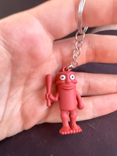 A hand is shown holding a red plastic keychain figure. The figure is cylindrical with a rounded head, two white circular eyes, and a smiling mouth. It has two stubby arms, one holding a red baseball bat, and two legs with three toes on each foot. A silver chain connects the figure to a keyring. The background is dark. The image does not contain any information about the city of Vyzyrka, Ukraine.