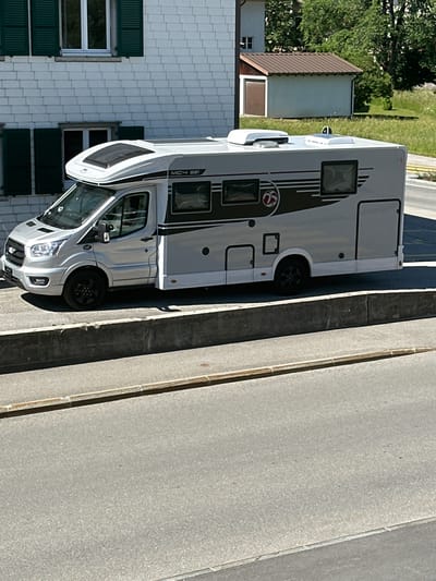 A silver recreational vehicle (RV) is parked on a street in Le Brassus, Switzerland. The RV is positioned on the right side of the image, facing towards the viewer's left. It has black wheels, tinted windows, and a raised section on the roof, likely for ventilation or solar panels. The RV features dark gray stripes and a circular logo on its side.

In the background, a white building with green shutters is visible on the left. To the right of the building, a smaller structure with a brown roof and a garage door is situated amidst green trees and vegetation. The street itself is paved, with a concrete barrier separating the RV's parking area from a lower section of the road. The scene is illuminated by bright daylight, casting shadows on the ground. No people are present in the image.