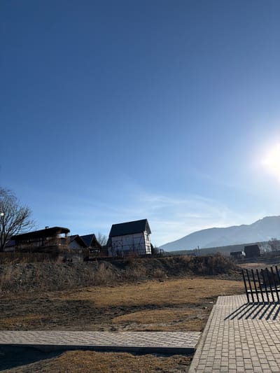 The image shows a landscape in the city of Manzherok, Russia. A partially obscured building with white facades and dark timber framing is visible. Adjacent to it are other buildings of similar construction, with dark rooflines. Behind the buildings, a mountain range rises, partially shrouded in a light haze. The foreground includes a dry, grassy slope and a paved brick walkway. A dark-colored railing casts shadows onto the walkway. Above, a clear blue sky with a bright sun.