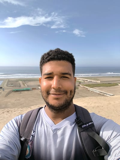 A selfie of a man with dark hair and a beard. He is wearing a grey and white athletic shirt and a black backpack. He is smiling. In the background is the Pacific Ocean and a strip of beach with what appears to be farming plots. The location is Huanchaco, Peru.