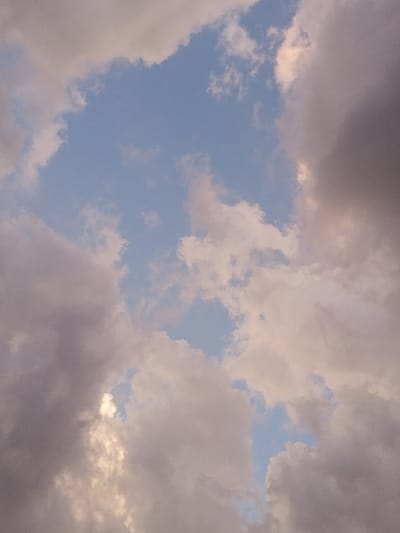 The image presents an upward view of the sky over New Koforidua, Ghana. The scene is dominated by a complex arrangement of clouds interspersed with irregular patches of clear, light blue sky. Cloud formations vary in density and color, ranging from lighter, off-white to pale grey cumulus-like clouds, some exhibiting a slight pinkish hue, to darker, denser grey masses. A notable area of brighter illumination, consistent with sunlight breaking through or reflecting off clouds, is visible towards the lower-left quadrant. No human subjects, animals, or man-made objects are present within the frame; the entire visual field consists solely of atmospheric elements.
