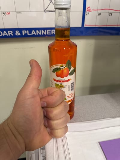 The image shows a person's thumb giving a thumbs-up gesture in front of a bottle of orange-colored liquid. The bottle has a label with images of fruit and text in English and Greek. Behind the bottle and thumb, a portion of a calendar is visible, with dates marked for November. The overall environment appears to be an indoor setting, possibly an office or a home, with a desk or table in the foreground. There are no discernible landmarks that would place this image specifically in the city of Kent, United States of America.