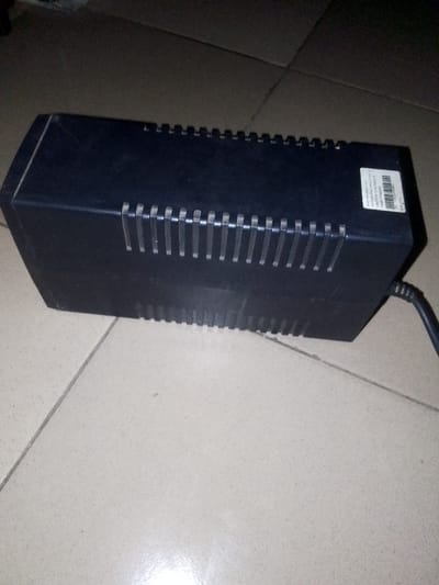 The image shows a black UPS unit lying on a tiled floor. The UPS has a series of ventilation slots and a barcode sticker on its top. It is connected to a black power cord, which is partially visible in the image. The location is Ushapa, Nigeria, which is a residential area with coordinates of latitude: 9.225, longitude: 7.41. The floor tiles are a light beige color and are arranged in a grid pattern. There are no other objects or people present in the image.