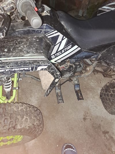 The image depicts a partially visible Telstar ATV in Obruchishte, Bulgaria. The ATV is dirty, with visible mud on the body and tires. The front left portion of the ATV is in focus, showing the handlebar, seat, engine, and front tire. The vehicle is parked on a concrete surface indoors. A single shoe is visible on the concrete floor near the right side of the image. The overall lighting is dim, and no people are visible in the frame.