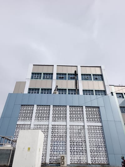An exterior low-angle shot shows the upper floors of a modern building with a light gray sky in the background. The building has a light blue facade on the lower section, featuring a large, decorative white lattice screen with a repeating geometric pattern. Above this, there is a section with light beige panels and several rows of windows with white frames. The windows are a mixture of open and closed. A black pipe runs vertically along the facade between some of the window sections, and an air conditioning unit is mounted to the wall. A white, rectangular concrete structure is situated in front of the lattice screen. In the lower left corner, part of an outdoor air conditioning unit is visible.