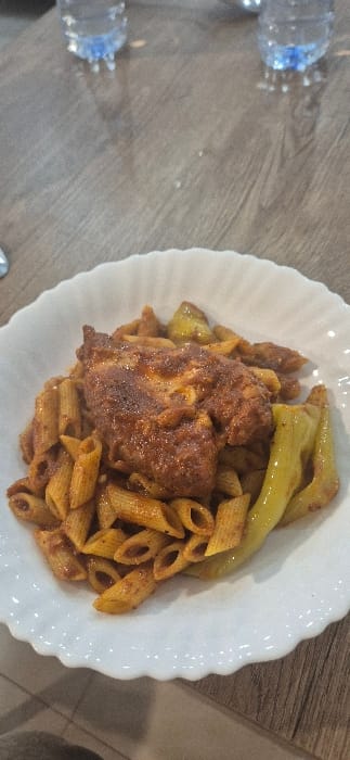A close-up, slightly overhead view displays a prepared meal on a light brown wood-grain table in Tixeraïne, Algeria. The central subject is a white ceramic plate with a scalloped rim, holding a serving of penne pasta coated in a reddish-orange sauce. Resting atop the pasta is a piece of reddish-brown, seasoned chicken. To the right of the chicken and pasta are several green, elongated vegetables. In the blurred upper background, two clear plastic water bottles with blue caps are visible. No individuals or active interactions are depicted within the frame.
