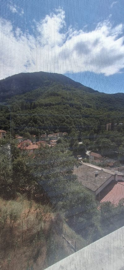 The image captures a view through a screen, looking out at a town nestled at the base of a densely forested mountain. The sky above is blue with scattered white clouds. In the foreground and midground, there are residential buildings with terracotta tile roofs, some partially obscured by trees. The mountain in the background is covered in lush green foliage, suggesting a rural or semi-rural setting. The screen's mesh pattern is visible across the entire image, creating a grid-like overlay. The scene appears to be in Нареченски бани, Bulgaria, a location known for its mountainous terrain and natural beauty.