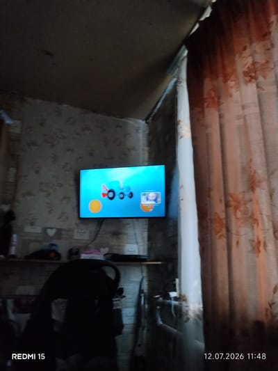 The image depicts an interior scene, likely a living room or bedroom, with a television screen prominently displaying a cartoon. The room appears somewhat cluttered and dimly lit. On the screen, a blue cartoon tractor is visible against a bright turquoise background. To the left of the tractor, a yellow circle is present. In the lower right corner of the television screen, a small inset shows a cartoon character with blonde hair and a smiling face.
The walls are covered in patterned wallpaper, and the ceiling appears to be made of rough material. To the right of the television, there is a translucent curtain with a floral pattern in shades of orange and brown. In the foreground, a dark silhouette of what appears to be a chair or stroller is visible on the left side of the image.
The bottom left corner of the image shows the text "REDMI 15", indicating the device used to capture the photo. The bottom right corner displays the date and time: "12.07.2026 11:48".
The location provided, Айет, Kazakhstan, is a remote village in the Aktobe region, known for its agricultural activities. While the image itself does not contain any direct visual cues to confirm this specific location, it presents a typical domestic setting that could be found in such a region.