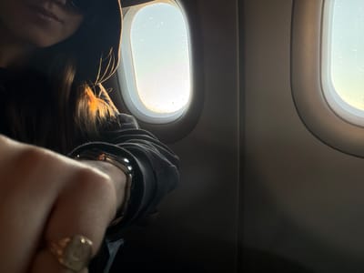 The image captures an intimate view inside an airplane cabin, focusing on a person seated by two windows. The central subject, likely a female, is seen from behind and slightly to the side, their head resting or leaning near the rightmost window. Their dark hair is dramatically backlit by the bright outdoor light, creating a radiant golden halo and highlighting individual strands. They are wearing dark clothing, possibly a hoodie or sweater.

In the upper right foreground, a hand is prominently visible, adorned with a golden signet ring and a metallic watch or bracelet. This hand partially frames the view of the person's upper body.

To the left, two oval airplane windows offer a bright, diffused view of the outside, which appears hazy or overcast. Both windows are covered with numerous small water droplets, indicating rain or condensation. The interior walls of the plane are a muted, light grey-brown. The overall lighting creates a high-contrast scene, with the bright windows contrasting sharply with the shadowed cabin interior and the person's figure.

The scene depicts a quiet moment during travel, possibly a flight over Lonate Pozzolo, Italy, as provided by the context. However, no specific external visual cues are present in the image to confirm this location. The strong backlighting suggests daytime. No discernible text is visible in the image.