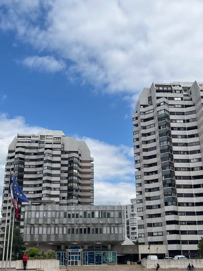 This media file shows a low-angle view of two large, modern apartment buildings against a bright blue sky with scattered white clouds. The buildings are tall and appear to be constructed from concrete and glass. Both have a repetitive, modular design with numerous windows. The building in the foreground has a grey facade and a distinctly zigzagging balcony structure. The building in the background, visible on the right, is also grey and has a more uniform facade with rows of windows.
In the lower right of the image, a few people are visible, including one person wearing a red jacket sitting on a ledge. There are also some parked vehicles, including what appears to be a white van. A flag, likely the European Union flag due to its blue color and yellow stars, is prominently displayed in the foreground, waving in the breeze. Other flags are also visible, suggesting a public space. Some signage is visible on the building in the background, but the text is too small to read clearly. The overall scene suggests daytime in an urban setting. The clear sky and bright lighting indicate fair weather. The architecture suggests a European location, consistent with the provided location context of Créteil, France.