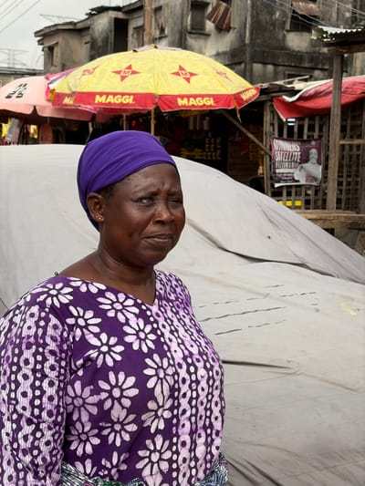 The image depicts a woman standing in an outdoor market setting within Jakande, Nigeria, under an overcast sky. She is positioned slightly off-center to the left, facing towards the right with a neutral expression. The woman has dark skin and wears a vibrant purple headwrap, a purple V-neck top with a white floral pattern, and the edge of a multi-colored patterned fabric is visible at her waist. Her left eye (viewer's right) exhibits a noticeable bluish tint. In the background, a bustling market environment is visible with several stalls and structures. A prominent yellow umbrella bearing multiple "MAGGI." logos in red provides shade over a stall displaying various packaged goods. To the left, a partial pink umbrella is visible. Behind these, distressed multi-story concrete buildings with visible wear, some featuring corrugated metal elements, form the urban backdrop. A large white tarpaulin-covered object occupies much of the middle ground behind the woman. To the far right, a poster is affixed to a wooden structure, partially obscured, with the legible text "WINNERS' SATELLITE FELLOWSHIP" and an image of a man. Utility lines are discernible in the upper left corner of the background.