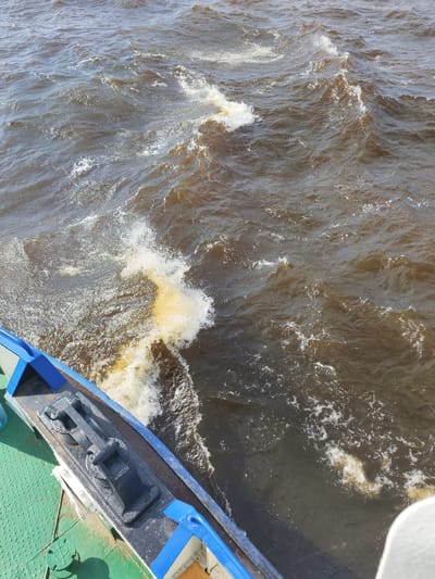 The image shows a view from a boat at the confluence of the Sheksna River and the Volga River in Krokhino, Russia. The water is choppy and brown, with foam and spray visible where the boat is moving. The edge of the boat, painted green and blue, is visible in the foreground, with a black cleat attached to its deck. The scene depicts movement through the water, suggesting the boat is in transit.