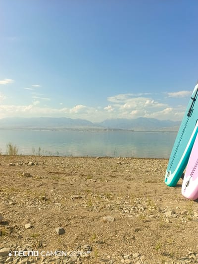 A wide shot captures a serene landscape with a large body of water stretching towards distant mountains under a bright blue sky with scattered clouds. In the foreground, a dry, rocky shore is visible, sparsely dotted with small green plants. To the right of the frame, two stand-up paddleboards, one teal and one pink, are leaning against an unseen object. The water reflects the sky and mountains, creating a calm and expansive scene. The horizon line is clearly defined, with the water occupying the lower half of the image and the sky the upper half. The overall impression is one of natural beauty and tranquility. The image also contains a watermark in the bottom left corner that reads "TECNO CAMON 20 Pro 5G".