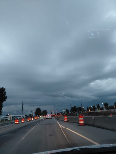 This media file captures a view from a vehicle traveling along a road at dusk, likely in Lexington, United States of America. The sky is heavily overcast with dark, dramatic clouds, suggesting an impending storm or the aftermath of one. The overall mood is somber and slightly tense due to the weather.

The scene is dominated by a roadway with construction barriers, indicated by numerous orange and white striped traffic barrels lining the side of the road. These barrels are a prominent feature, suggesting ongoing road work or maintenance. The road itself appears wet, reflecting the dim light, further emphasizing the weather conditions.

On the left side of the frame, a digital display is visible, partially obscured by the overcast sky. It shows some text and graphics, though it's not entirely legible. Further to the left, faint reflections can be seen on what appears to be a window or mirror.

On the right side of the road, there are trees and buildings, as well as utility poles with power lines. The lighting conditions indicate it is either early morning or late evening, with the artificial lights from street lamps and traffic lights starting to become more noticeable. The presence of traffic signals and other vehicles further down the road confirms this is an active roadway. No people are clearly visible, but the context implies the presence of drivers and potentially construction workers. The time of day is clearly dusk, with the sky a deep grey and the light fading.