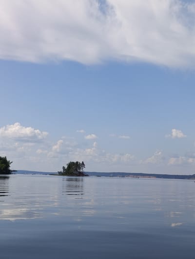 The image depicts a tranquil body of water, likely a lake or wide river, on a clear day with scattered white clouds in a blue sky. In the middle distance, a small island covered in dense green trees is visible. The water's surface is calm and reflective, mirroring the sky and clouds. The distant shoreline is low-lying and appears to be covered in vegetation. The scene is serene and natural, suggesting a rural or semi-rural setting. The location is identified as being in the city of Vekoshinka, Russia.