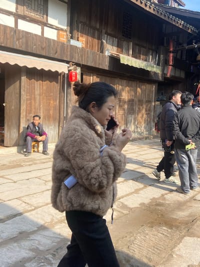 A woman in a furry brown coat talks on the phone on a stone path. Black pants contrast with the coat. Behind her is an aged, wooden building with red lanterns. Further back, two men are walking, one appearing to hold a phone. In the building's doorway, an elderly man sits on a stool, his hands clasped in his lap. The area appears sunlit and historical.
