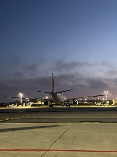 An airplane, identified by the tail logo as Air Portugal, is parked on the tarmac of the Porto Airport in Moreira, Portugal. The image captures the aircraft from a rear-facing perspective, with its engines clearly visible. The scene is set during the twilight hours, as indicated by the gradient of the sky, transitioning from a deep blue at the top to lighter shades with a hint of purple near the horizon. Clouds are present in the sky, obscuring some of the celestial view. The tarmac is illuminated by various lights, including runway lights and lights from the airport infrastructure. Red and yellow lines mark the taxiways on the concrete surface. Several airport support vehicles and equipment are positioned around the aircraft. The overall atmosphere is calm and still, characteristic of an airport at dusk or dawn.