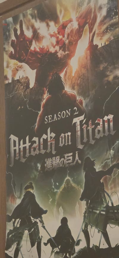 The image is a poster for "Attack on Titan Season 2". It features a large, monstrous titan in the upper portion, with a rocky, mountainous landscape and a fiery glow behind it. Below the titan, the text "SEASON 2" is prominently displayed, followed by the title "Attack on Titan" in large, stylized English lettering, and then Japanese characters below that. The lower part of the poster shows silhouetted figures of characters from the anime, some in action poses with swords. There is no indication of any location in the city of Shomron Regional Council, Palestinian Territories.