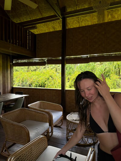 A woman with wet, dark hair, wearing a black bikini top, is standing in a bamboo-constructed building with open window views of lush greenery, including trees and red flowering plants. She is smiling, with one hand touching her head. The building features wicker furniture, including chairs with white cushions and a small round table. The location appears to be Santa Monica, Philippines.