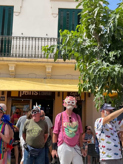 In Sitges, Spain, under a bright sun, a group of people are gathered on a street in front of a building with a yellow awning. The building has dark green shutters on the upper floor and a balcony with an ornate metal railing. The awning covers the entrance to a shop labeled "pâris & COMPANY".
Several individuals are prominently featured, some dressed in colorful and festive attire, suggesting a celebration. One man in the foreground wears a pink t-shirt with a kiwi fruit graphic, white pants, and pink hair rollers in his hair, along with novelty glasses. To his right, another man sports a green t-shirt with the text "THE NORTH FACE NO DAYS OFF NEVER". He is also wearing a fanny pack and a unicorn headband with sunglasses. Another man on the far right wears a white t-shirt printed with colorful toy cars. The people appear to be interacting with each other and engaging with the event taking place. The scene is lively, with a sense of community and gaiety.