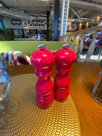 Two red ceramic salt and pepper grinders with chrome tops sit on a gold woven placemat on a wooden table. The grinders are positioned side-by-side in the foreground. To the left of the grinders, a portion of a clear glass is visible. In the background, a restaurant interior is apparent with green upholstered seating, colorful striped pillows, and decorative triangular patterns on a white wall. The restaurant appears to be in the city of Solnechnoye, Russia.