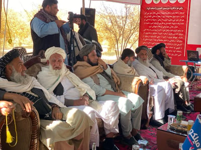 The image shows a group of men, possibly elders, sitting on ornate couches in an outdoor setting. They are dressed in traditional clothing, turbans and shawls. A man stands behind them, possibly recording or photographing the event. A banner with text in Arabic script is visible in the background, along with a landscape of trees and open space. A table with refreshments is placed in front of the group.  A logo with "Milko" is visible.