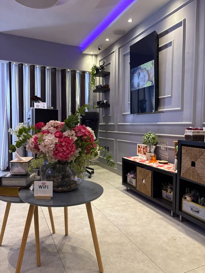 The image depicts an indoor scene, likely a reception area or waiting room in a salon or spa, given the aesthetic. The setting is modern and clean, with a gray tiled floor and white paneled walls. A large television screen is mounted on the wall, displaying a close-up of a woman's face with yellow flowers around her head. To the right of the television is a black shelving unit with woven baskets and decorative items, including small framed pictures and plants.
In the foreground, a round gray side table with wooden legs holds a large glass vase filled with a vibrant bouquet of pink and white hydrangeas and other greenery. Next to the vase, a small white sign with a QR code and the word "WiFi" is visible. There are also some decorative books and a tissue box on the table.
Behind the table, a staircase with dark wooden steps and illuminated edges leads upwards, suggesting a multi-level space. The lighting in the room is a mix of ambient and accent lighting, with a strip of purple light visible along the ceiling on the left side of the image. The overall mood is calm and inviting. There are no discernible cues about the time of day or weather, but the interior setting suggests it is not outdoors.
Visible text includes "WiFi" on the sign and text on a framed item on the shelf which appears to be a label for "GRAND JARDIN BULBES". There are also some smaller text elements on the QR code sign and on the television screen, but they are not legible.
