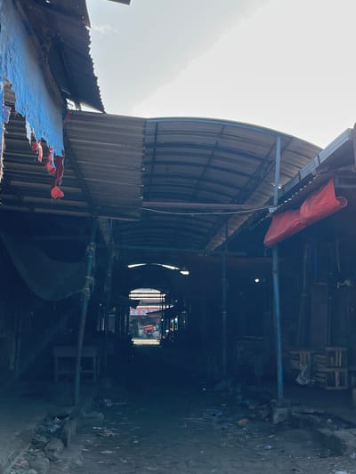The image depicts a dark, narrow alleyway or market corridor with a corrugated metal roof supported by metal poles. The roof is partially covered with blue and red tarps, some of which are tattered and hanging. The ground is uneven and covered with debris, including trash, rocks, and dirt. On the left side, there are some empty tables or stalls. On the right, there are stacks of wooden crates. In the distance, the alley opens up to a brighter area where a red vehicle, possibly a cart or small truck, is visible. The overall impression is one of a poorly maintained, possibly abandoned, market or service area in Juwet Kenongo, Indonesia. The lighting suggests it is daytime, but the area is heavily shaded.
