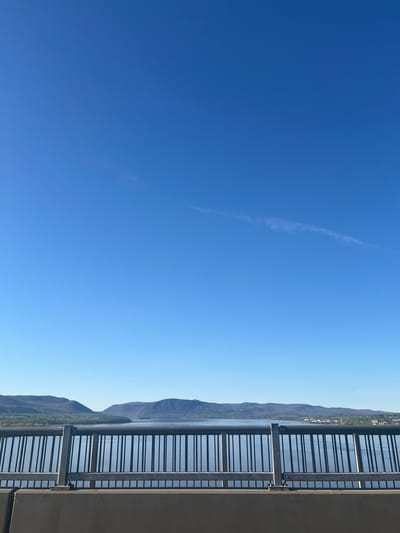 The image shows a view from a bridge or elevated platform, likely in the City of Newburgh, United States of America, given the presence of a waterfront, hills, and possibly a small town in the background. The primary focus is on a metal railing along the right side of the frame. Behind the railing, there is water and a mountainous or hilly landscape covered with some greenery. In the distance, a cluster of buildings suggests a town. The left side of the image is predominantly a clear blue sky with a faint contrail. The lighting is bright and suggests a sunny day.