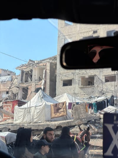 A damaged building, likely from conflict, dominates the view from inside a vehicle. The structure has visible holes and extensive debris. Makeshift coverings of white tarp are stretched across damaged areas. Clothes hang on lines strung between buildings. A group of people, including a man and a child, stand on the side. A dark object resembling a rearview mirror is visible in the foreground of the picture.