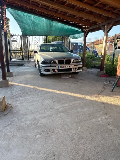 The image depicts a silver BMW sedan parked under a wooden carport with a green shade cloth. The carport has a tiled roof and concrete pillars. In the background, there is a metal fence, some vegetation, and residential buildings. A blue tractor is visible behind the fence. On the ground, near the car, are a broom and a pair of shoes. The license plate of the car reads "PB 0955 XT". The scene appears to be in a residential area in the city of Vinitsa, Bulgaria. The lighting suggests it is daytime, likely late afternoon, given the long shadows cast on the concrete ground.