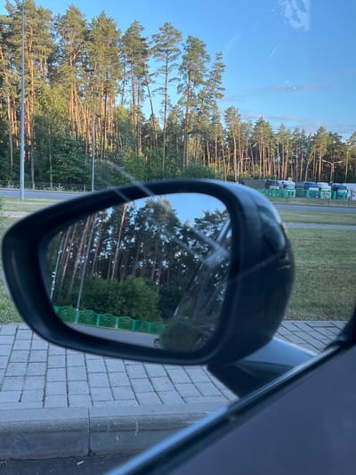 A side-view mirror reflects a dense forest of tall, slender pine trees against a clear blue sky. The mirror is framed by a black casing, and part of a dark car's interior is visible in the lower right corner. Beyond the reflection, the actual scene shows the same forest line, with a road and a green fence in the middle ground. Several trucks are parked in a lot behind the fence. The foreground features a paved walkway with square gray bricks. The image appears to be taken from a vehicle, suggesting a roadside or parking area. The location is identified as Babovichi Village Council, Belarus.